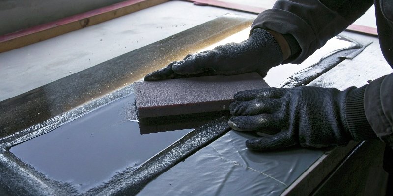 A person wearing thick gloves manually sanding a glass edge with wet sandpaper on a block